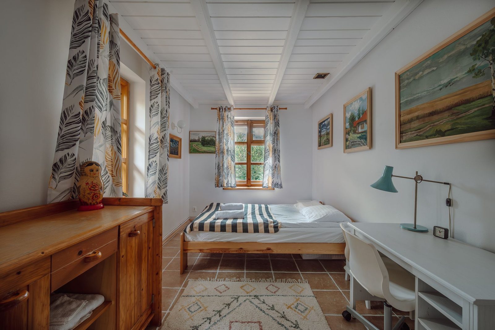 Bright bedroom with a single bed, paintings on the wall and a desk by the window